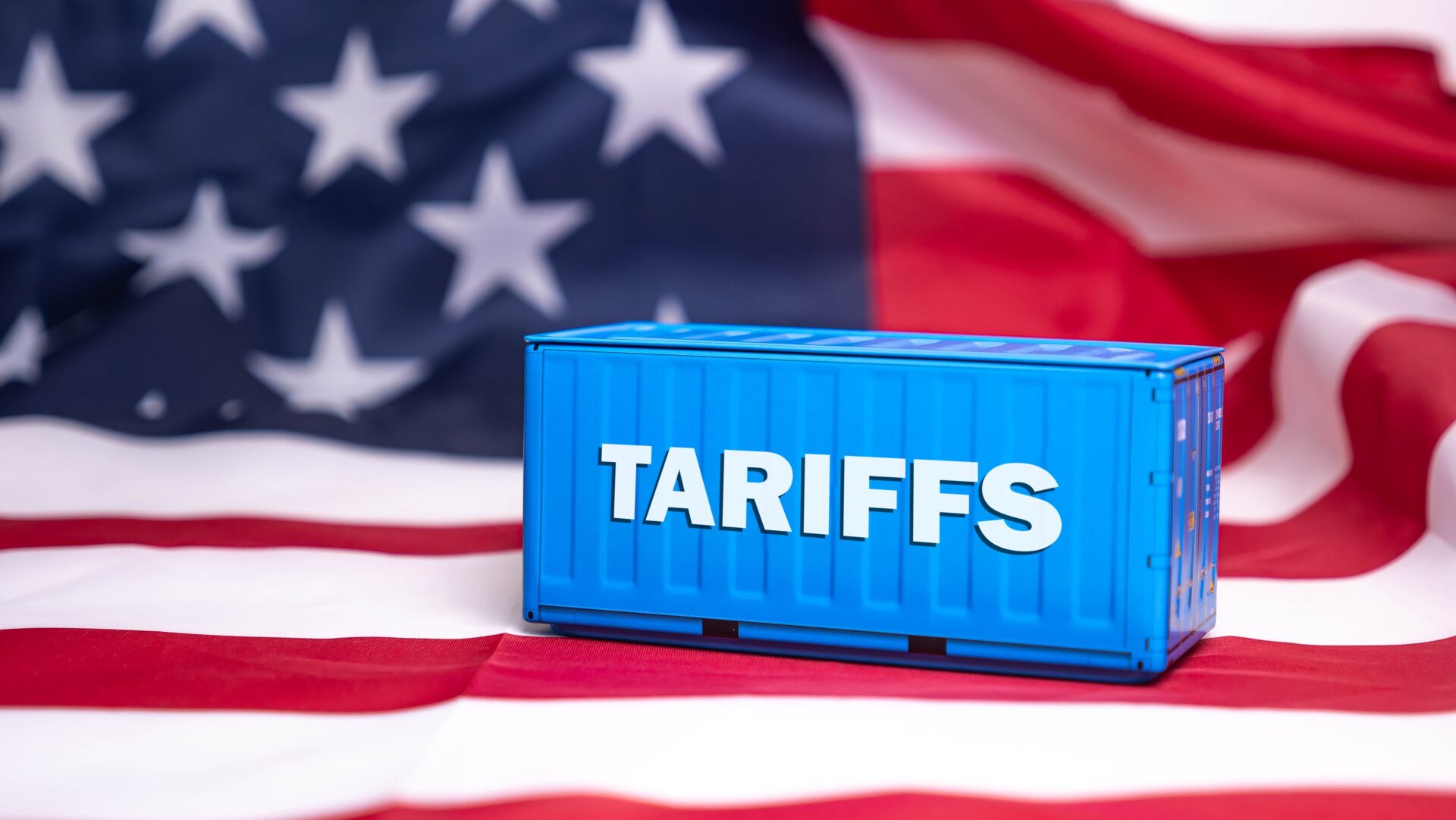 Amerikanische Flagge im Hintergrund, darauf ein Seecontainer mit dem Schriftzug "Tariffs"